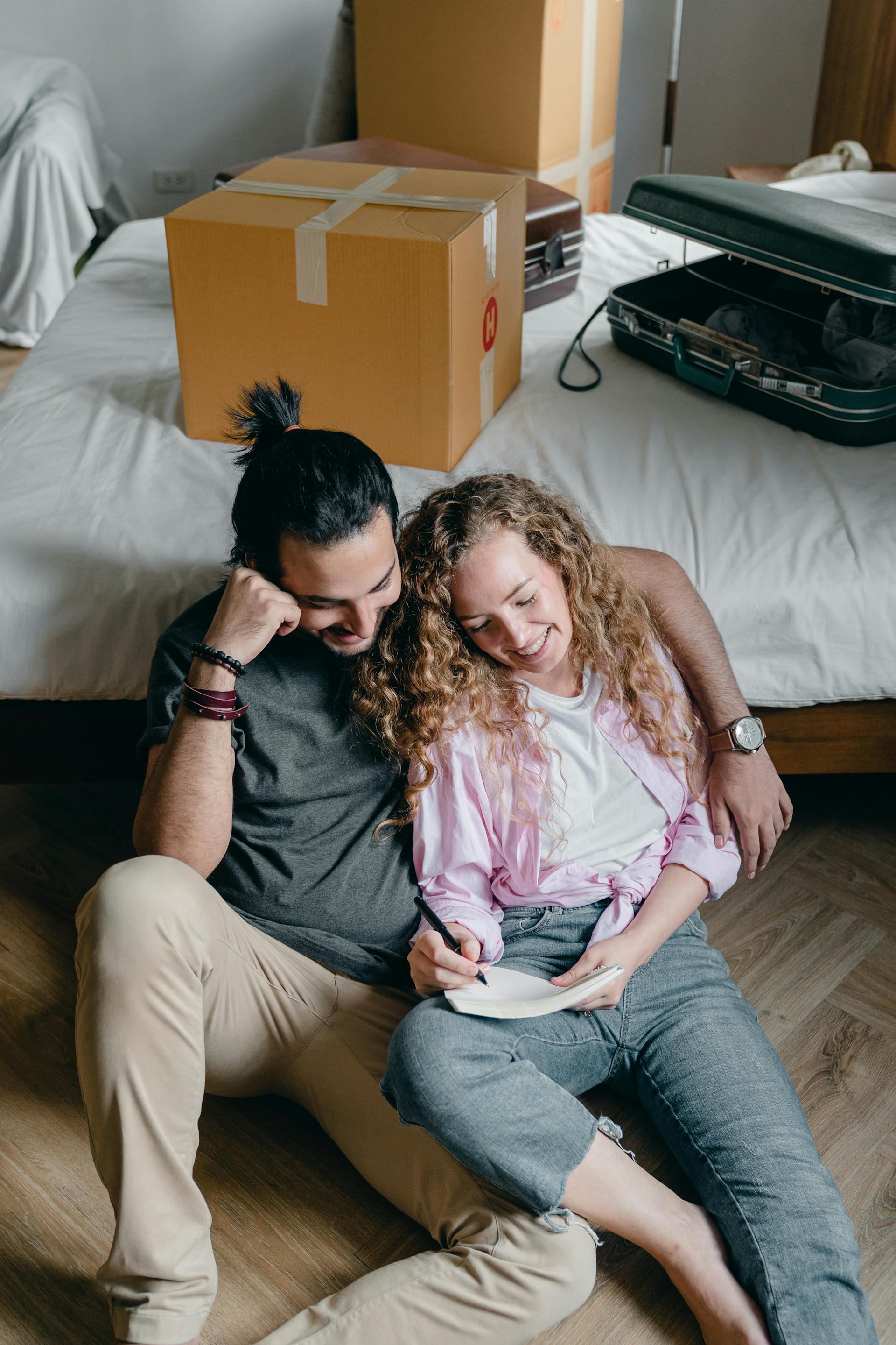 A happy couple sits on the floor amidst moving boxes, leaning against the bed. The man, with dark hair tied in a top knot, has his arm around the woman, who is smiling and writing in a notebook.