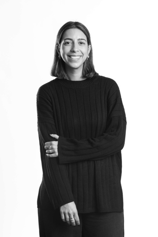 Black-and-white studio portrait of a smiling woman with shoulder-length straight dark hair, wearing a black crewneck sweater and rings, with one arm crossed over the other, against a plain light background.