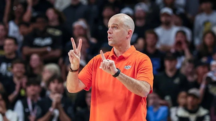 A basketball coach in an orange polo shirt stands courtside, raising his hands to signal plays with his fingers during a game. A blurred crowd is visible in the background.
