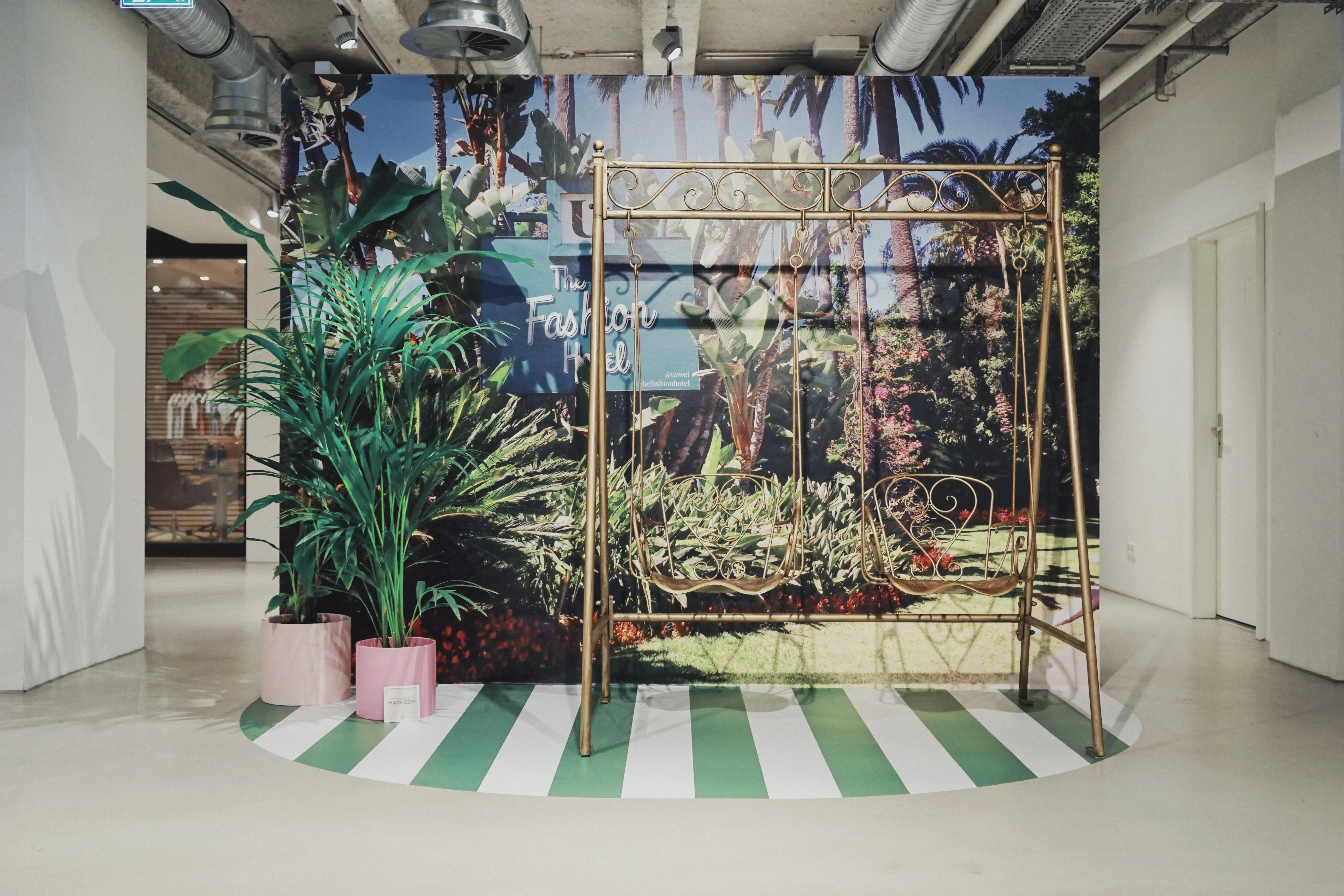 Photo-booth corner at UZWEI FASHION HOTEL with a golden swing bench in front of a large palm-garden mural with The Fashion Hotel sign, pink planters and a green-striped floor circle.