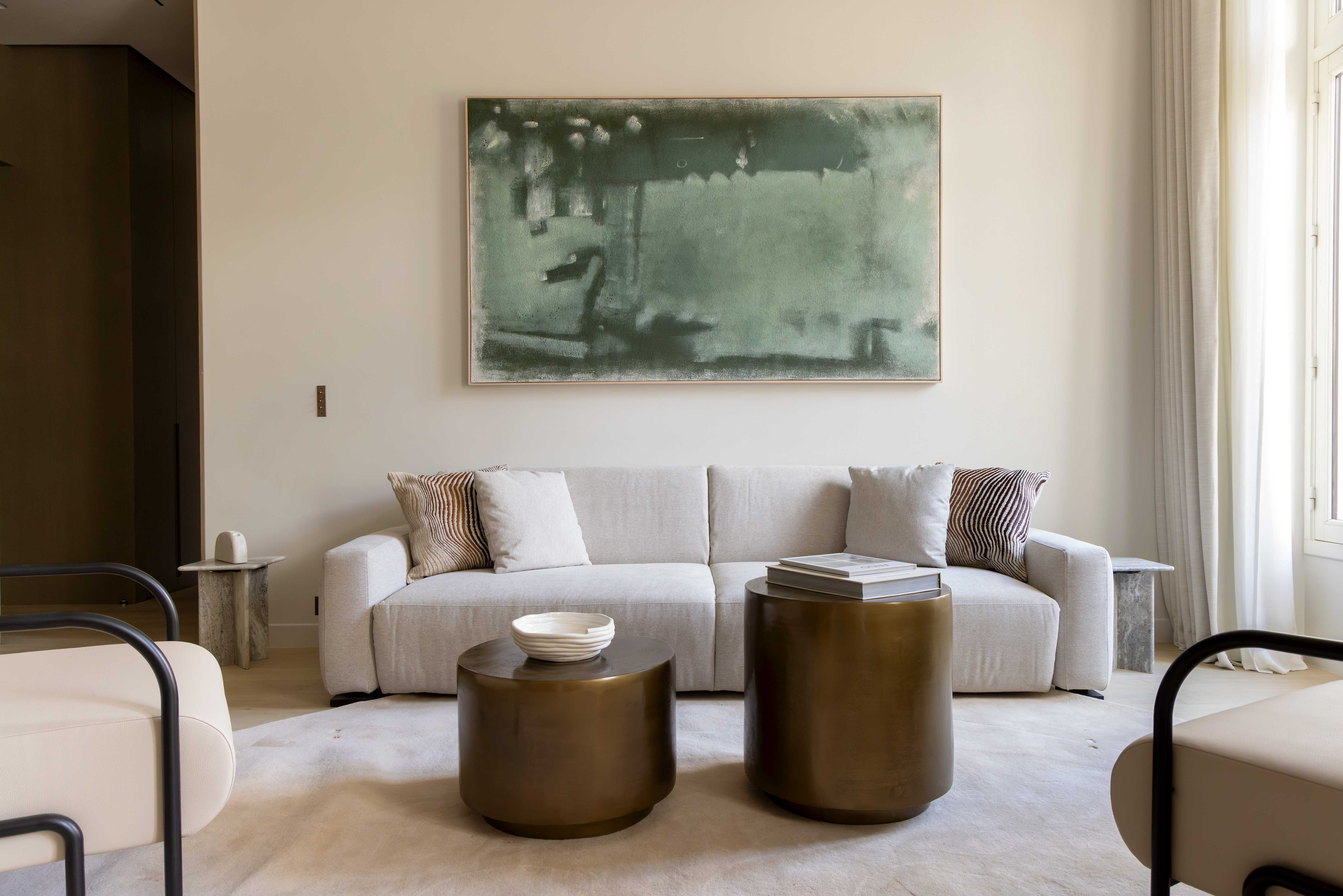 Canape beige avec coussins, grande toile abstraite verte au mur, tables basses cylindriques en laiton