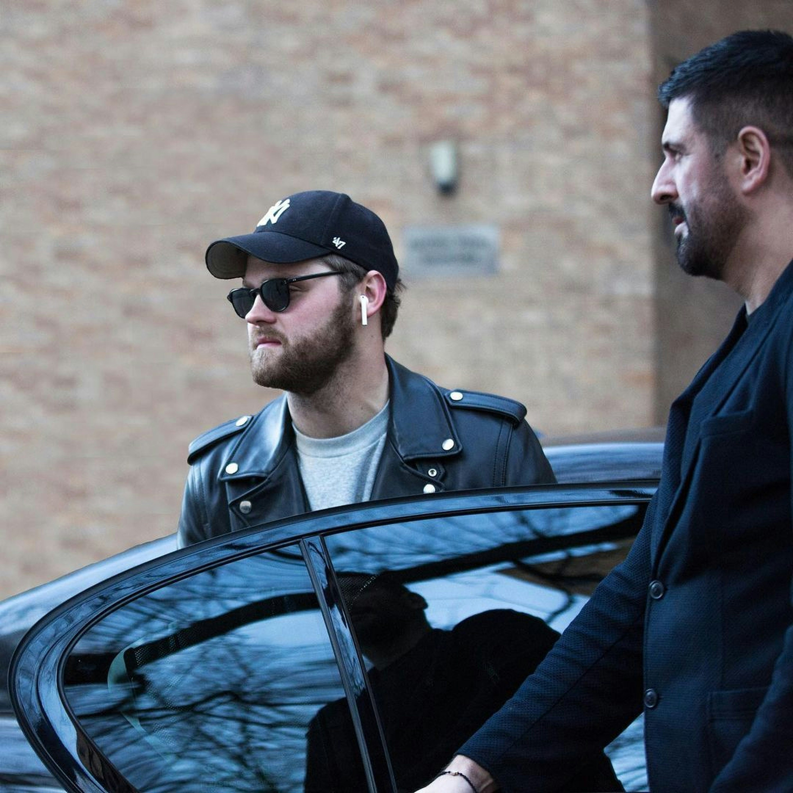 Man in leather jacket exits car with security
