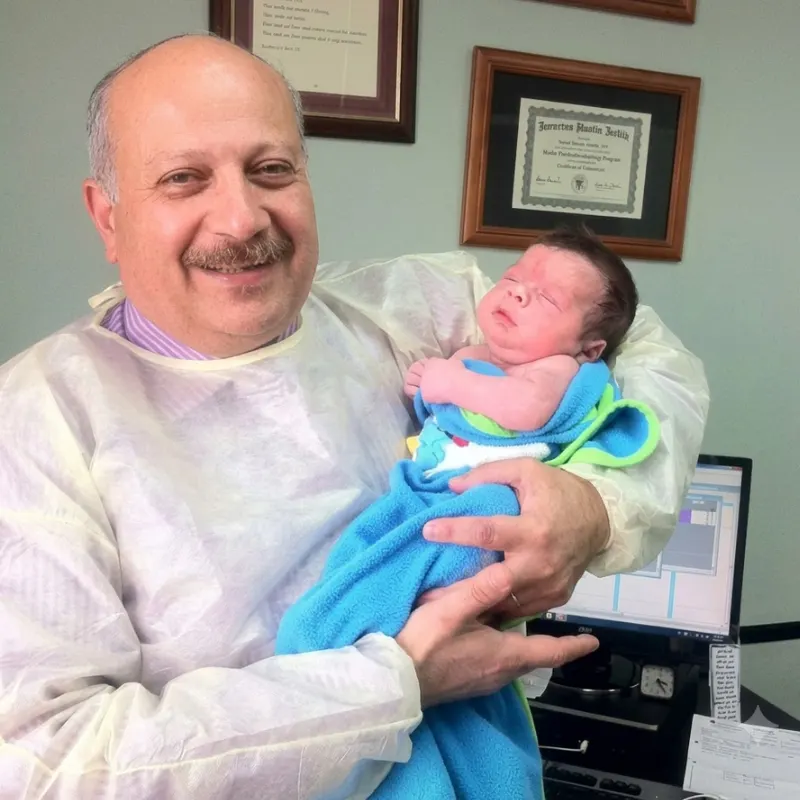 Dr. Ashraf Saddik cradling a sleeping newborn baby wrapped in a blue blanket in front of framed medical certificates at his Coburg clinic.