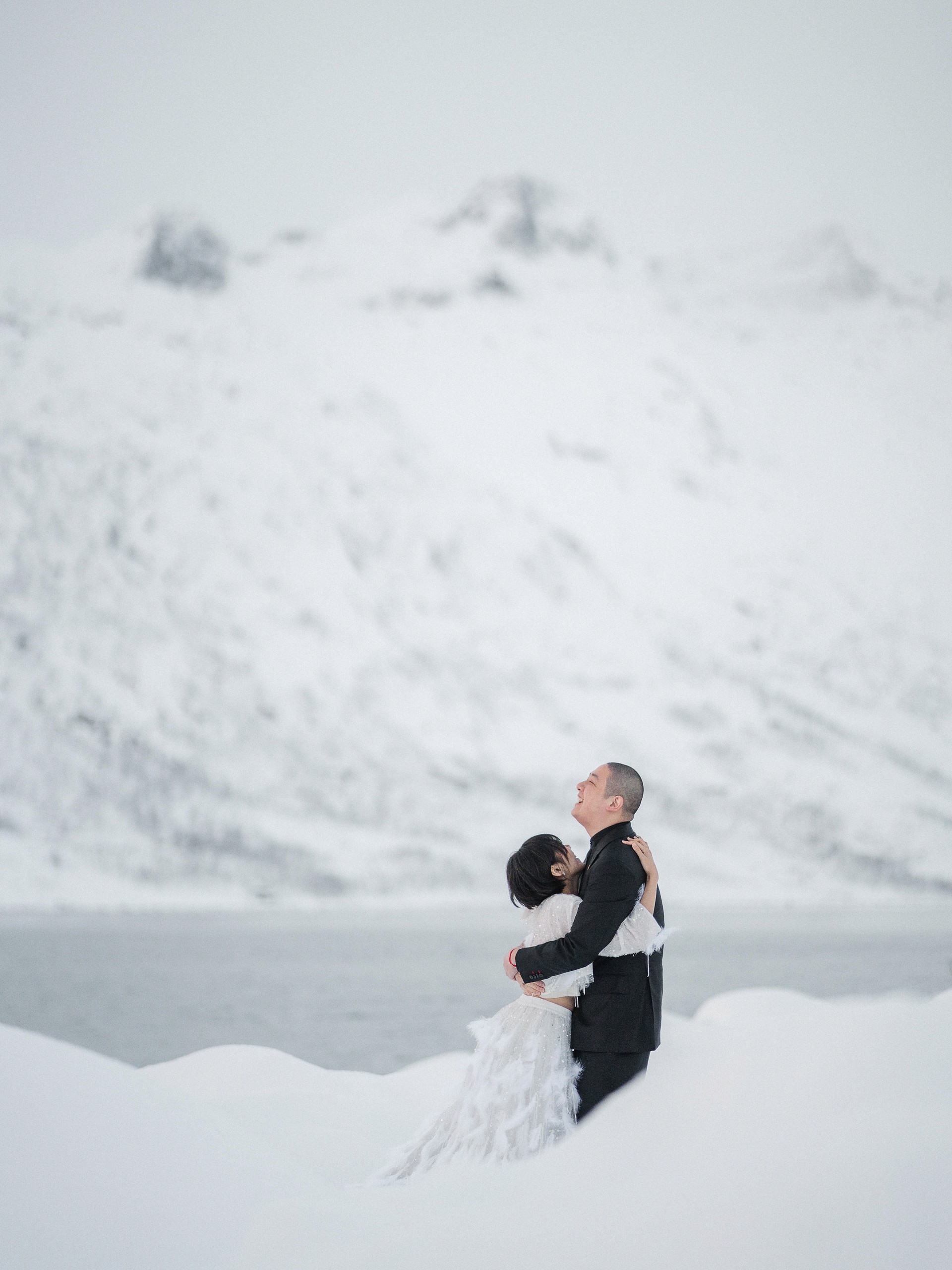 The couple appears in profile within a vast white snowfield, the groom looking upward as the bride leans into him beside the water.
