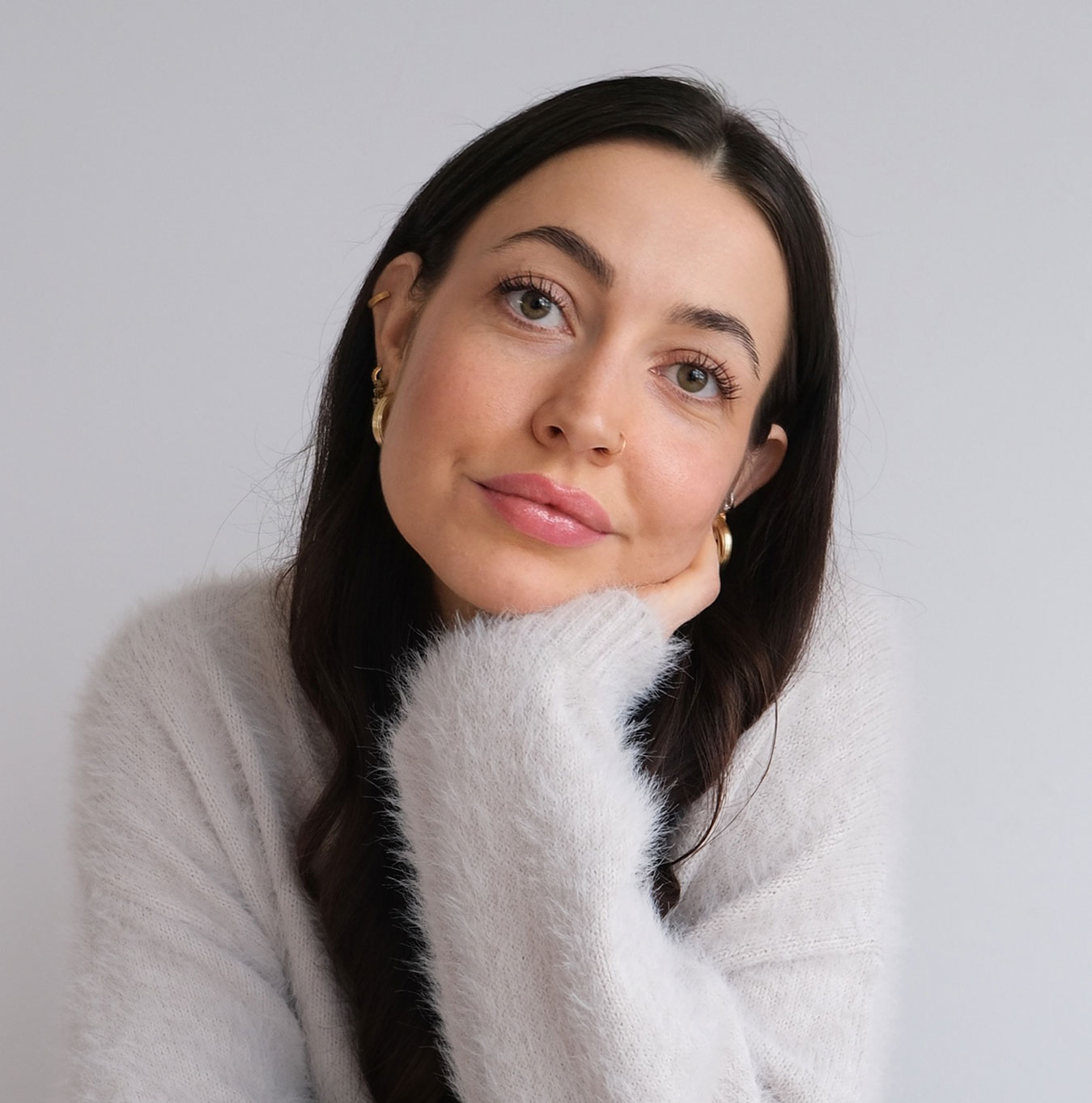 Minimal studio portrait of a confident woman in a white shirt, arms crossed, on a soft gray background.