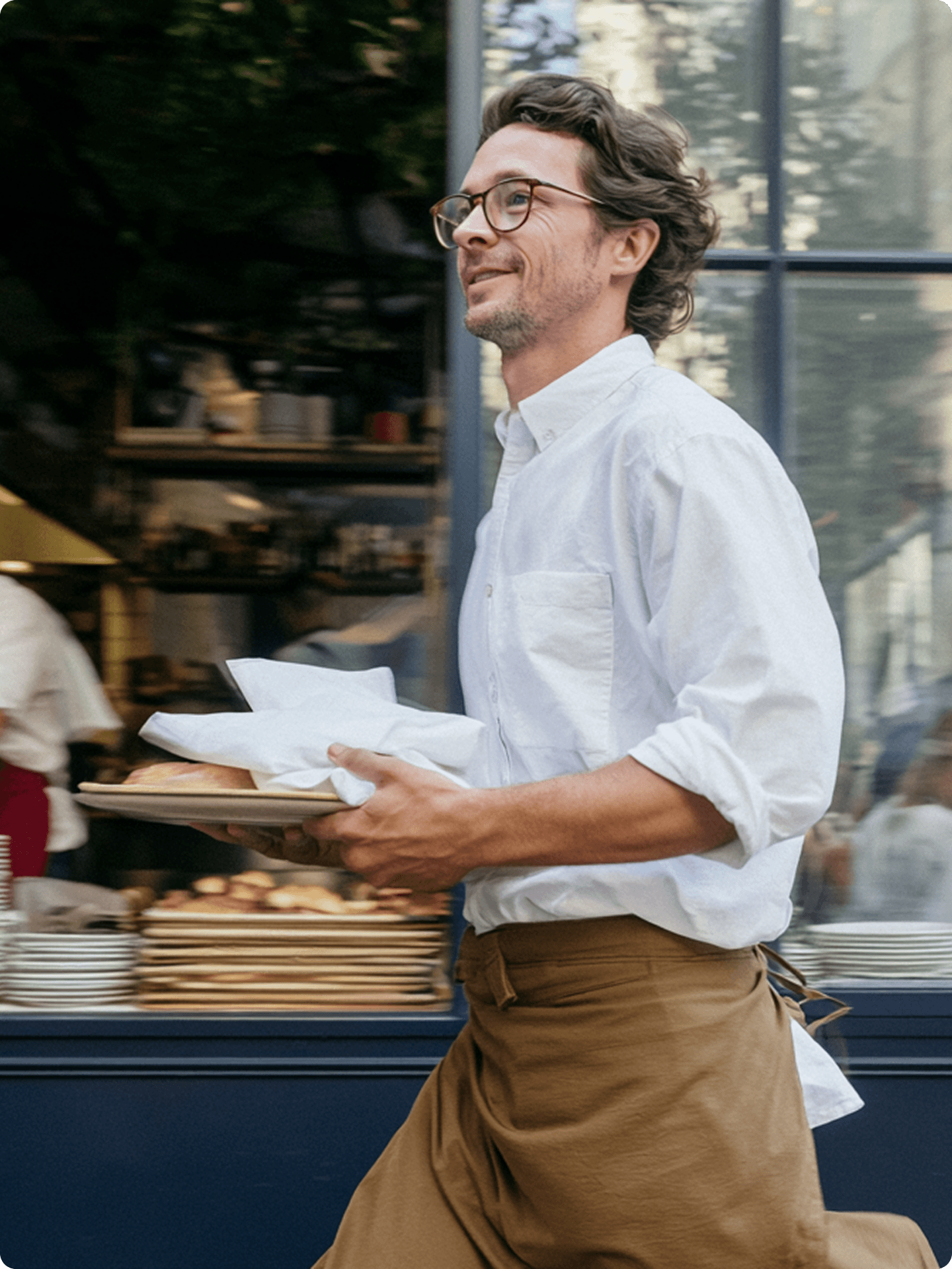 Man in a white shirt and tan apron carrying a tray of wrapped bread past a bakery window.