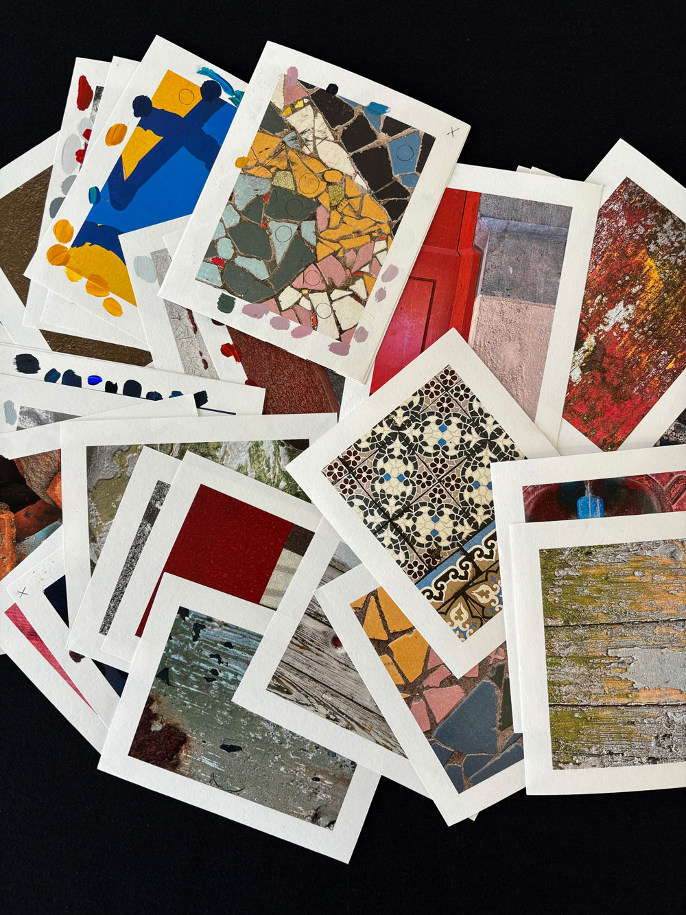 Een bovenaanzicht van een chaotische stapel witte kaarten op een zwarte ondergrond. Elke kaart toont een close-up foto van texturen of patronen, zoals mozaïek, verweerd hout, tegels en abstracte verfstreken in verzadigde kleuren.