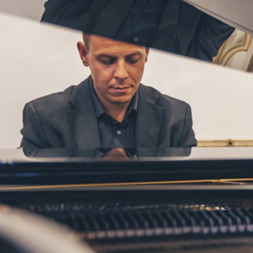 Man in a dark suit, seen through the open lid of a piano, focused on playing the instrument indoors.