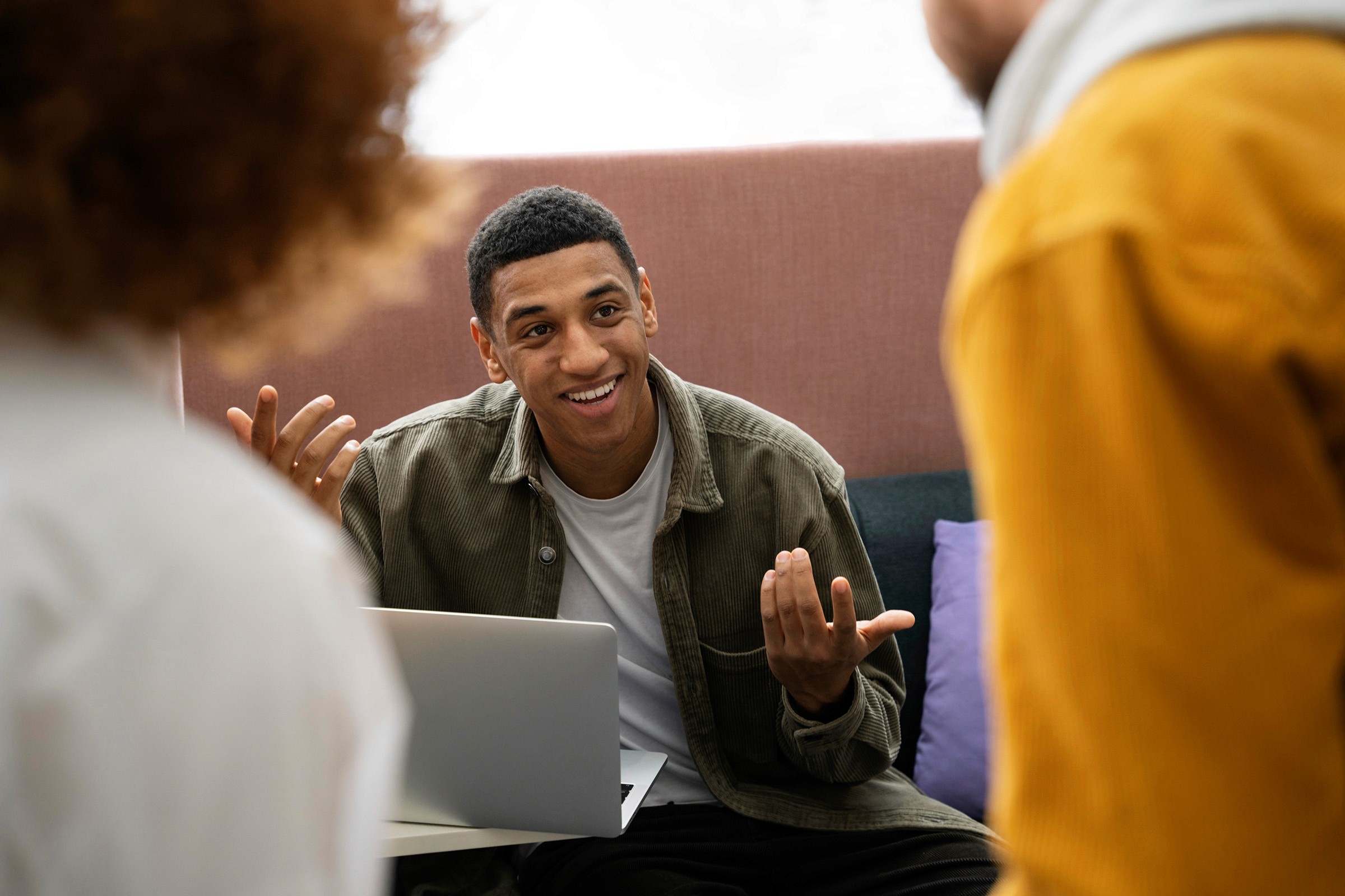 A smiling man engaged in a discussion with colleagues, gesturing while seated with a laptop in front of him