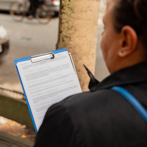 Person holding a blue clipboard with paper, reading on a street with parked cars and a bike in the blurred background.
