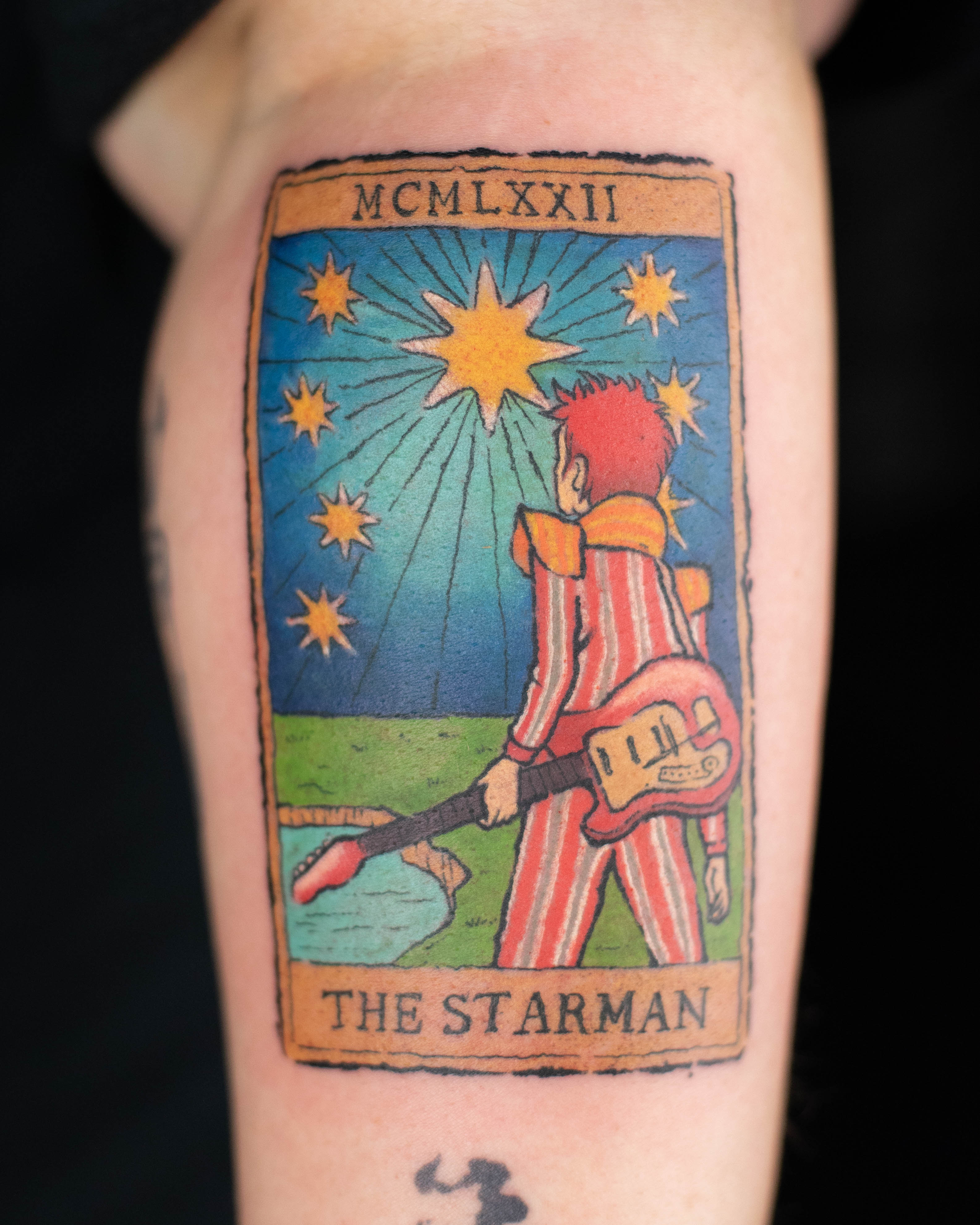 Full-colour tattoo styled as a vintage tarot card featuring Ziggy Stardust under a radiant starfield. The figure holds an electric guitar and wears a red-striped suit, captioned “THE STARMAN” with Roman numerals MCMLXXII. Tattoo by Samma Charles Art in Boston, MA and Oxford, UK.