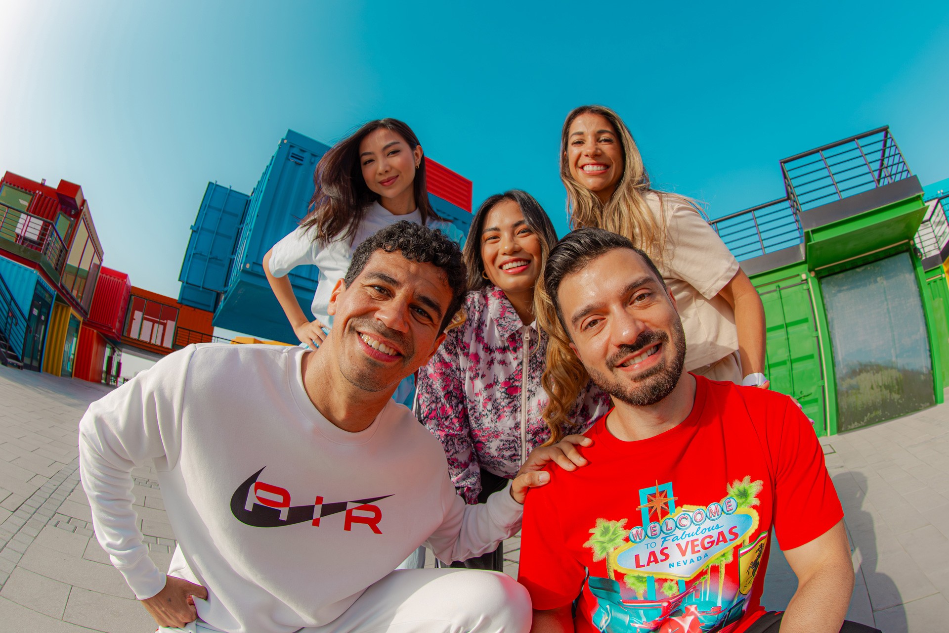 Five friends grouped against bright shipping containers, shot wide on a fisheye lens that curves the frame.