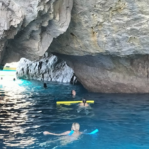 People swimming in a cave with clear blue water and natural rock formations above and around them. Some use floating devices.