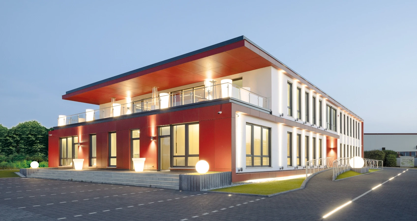 Modernes Bürogebäude mit roten Verkleidungen, Glasfronten und einem flachen Dach in Rottönen, beleuchtet am Abend.