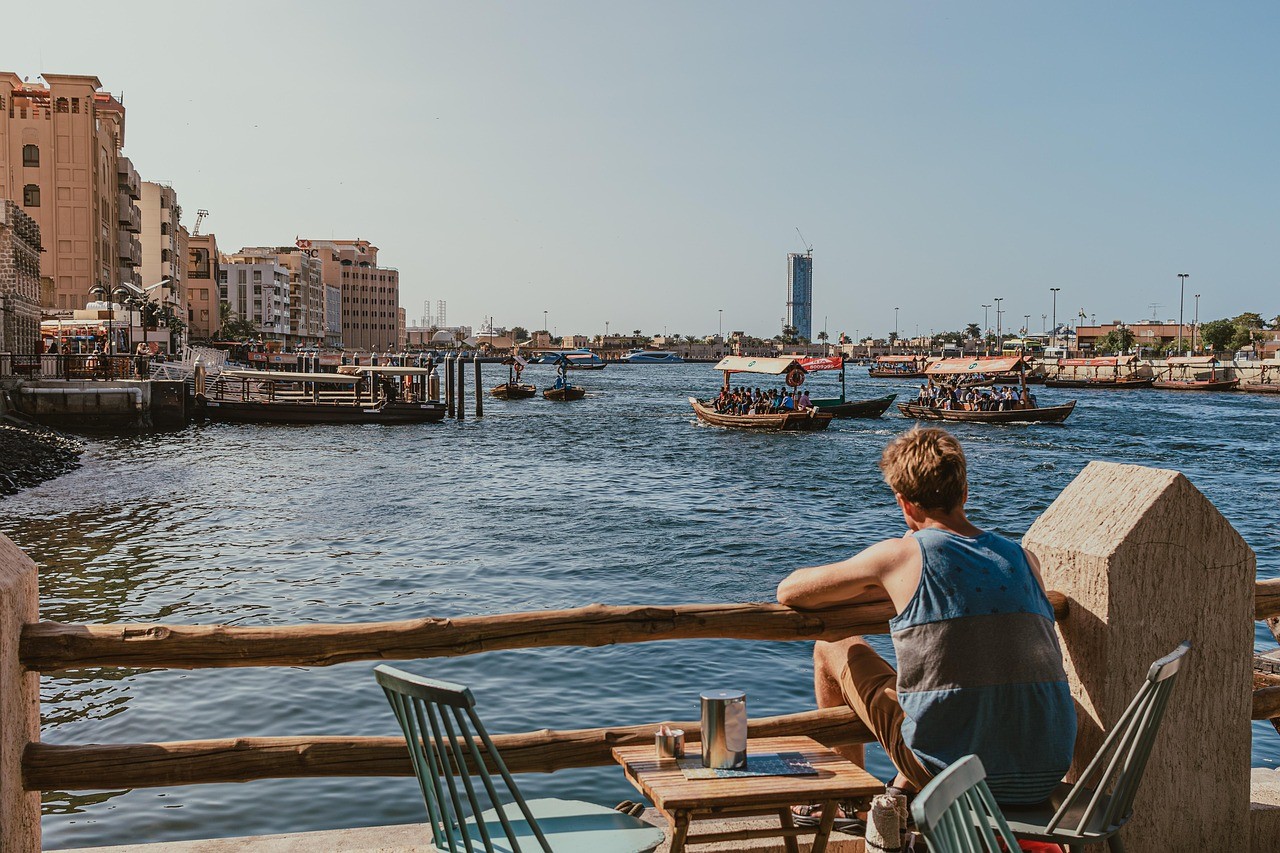 A man sits along Dubai Creek, with traditional abra boats and buildings along the shore.&nbsp;