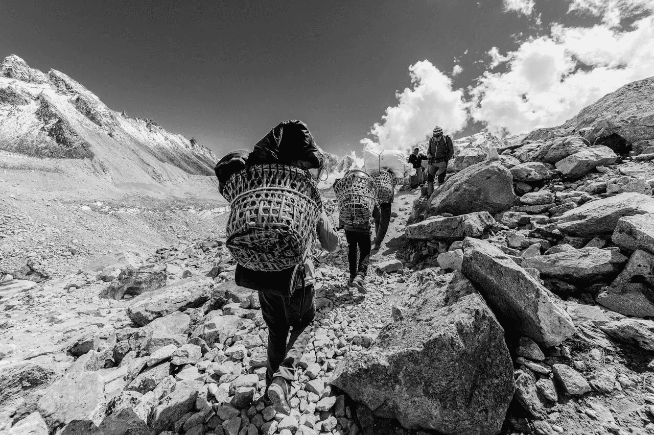 Black and white photograph of Sherpa guides carrying traditional dokos through a Himalayan mountain pass, representing the spirit and heritage behind Café Sherpa's in Brookfield, WI. A café built from years of travel across cultures and continents.
