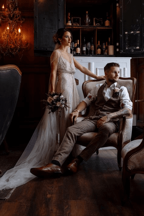 Stylish couple in a vintage-inspired setting, with the bride holding a bouquet and the groom seated in a classic armchair, exuding an old-world charm typical of vintage wedding photography.