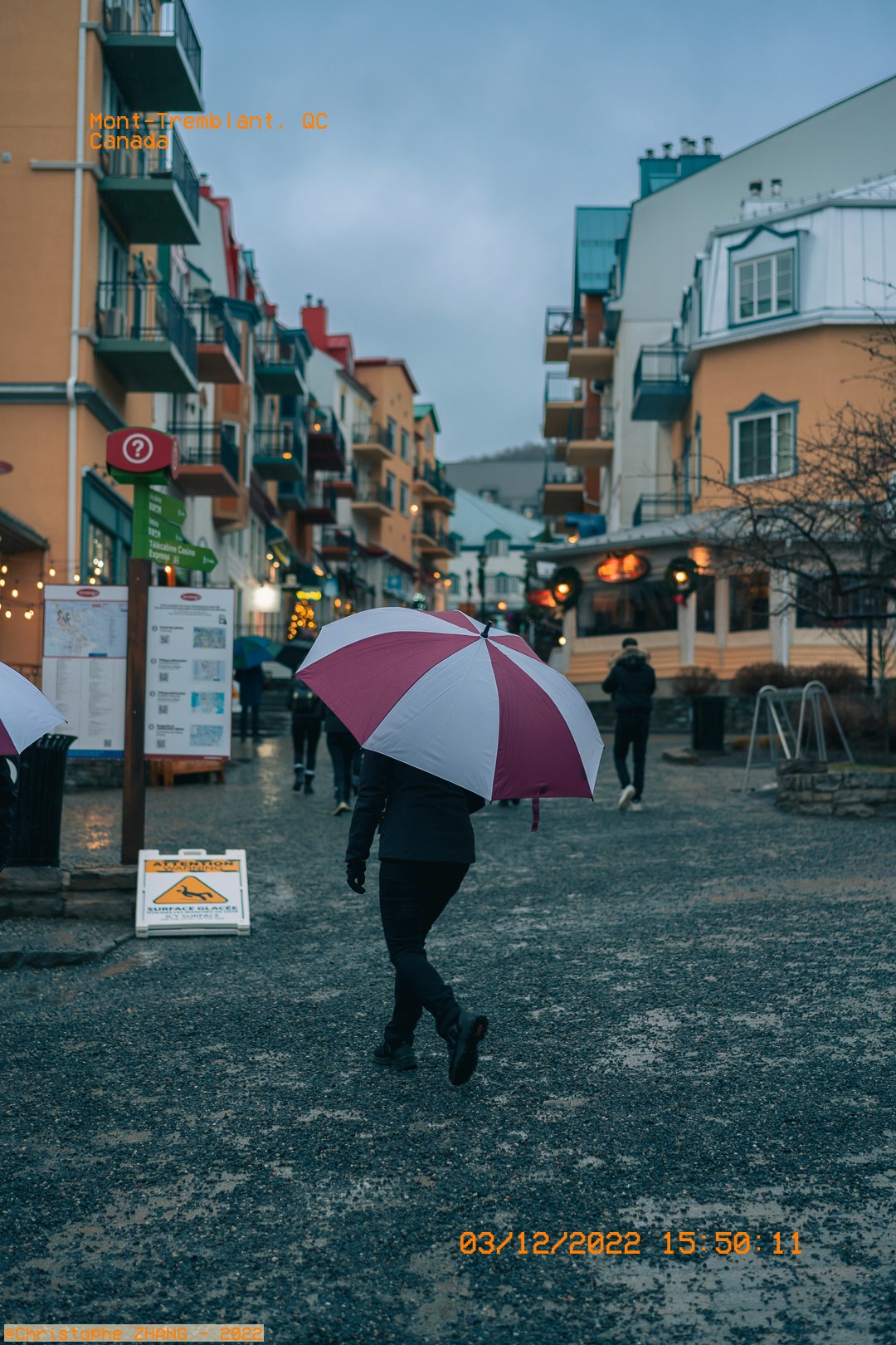 Promeneur avec un parapluie rose et blanc dans une rue de village sous la pluie.