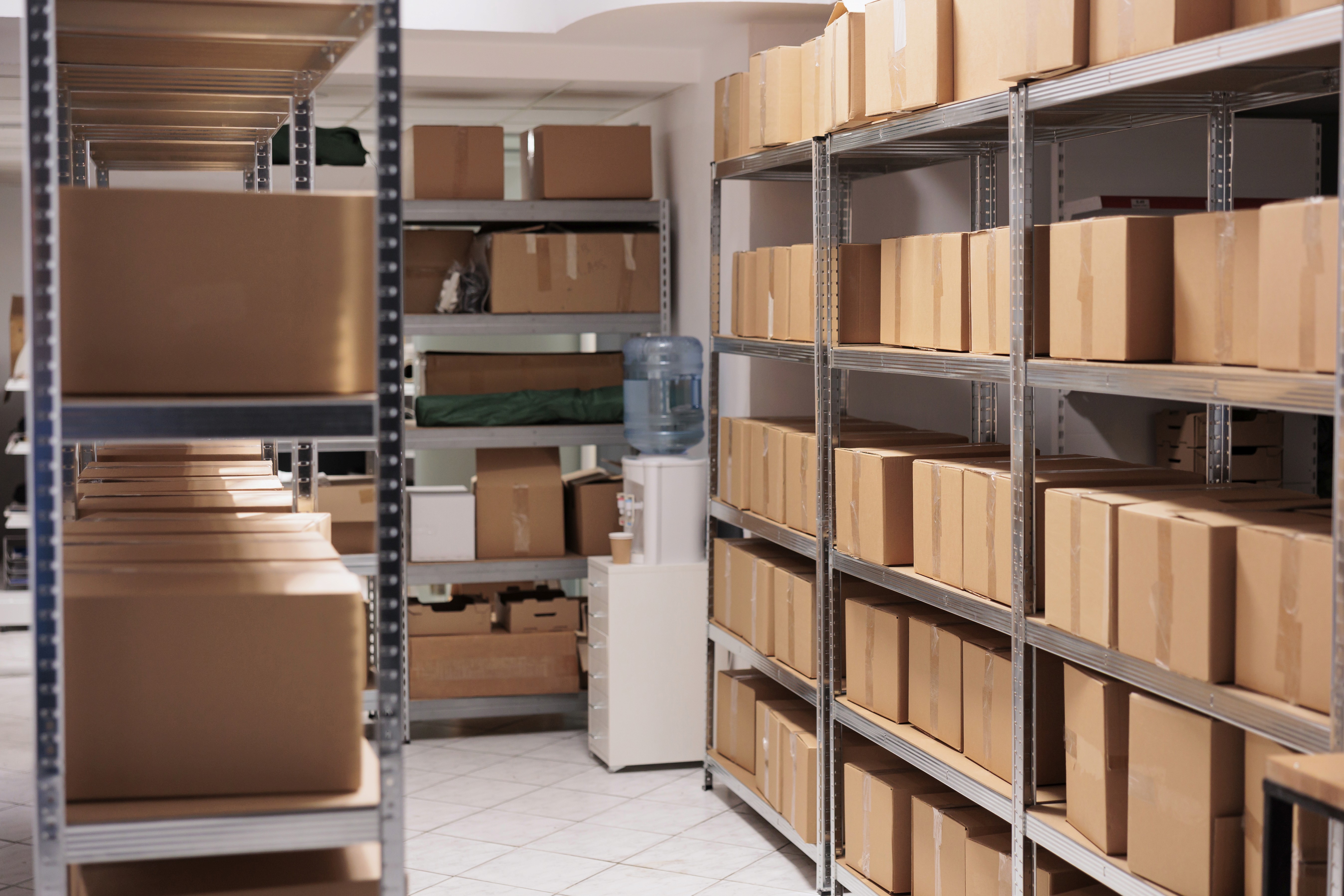Storage room with shelves filled with brown cardboard boxes in a well-organised storage room dedicated for business. A water cooler is visible in the background, creating a neat, orderly atmosphere.