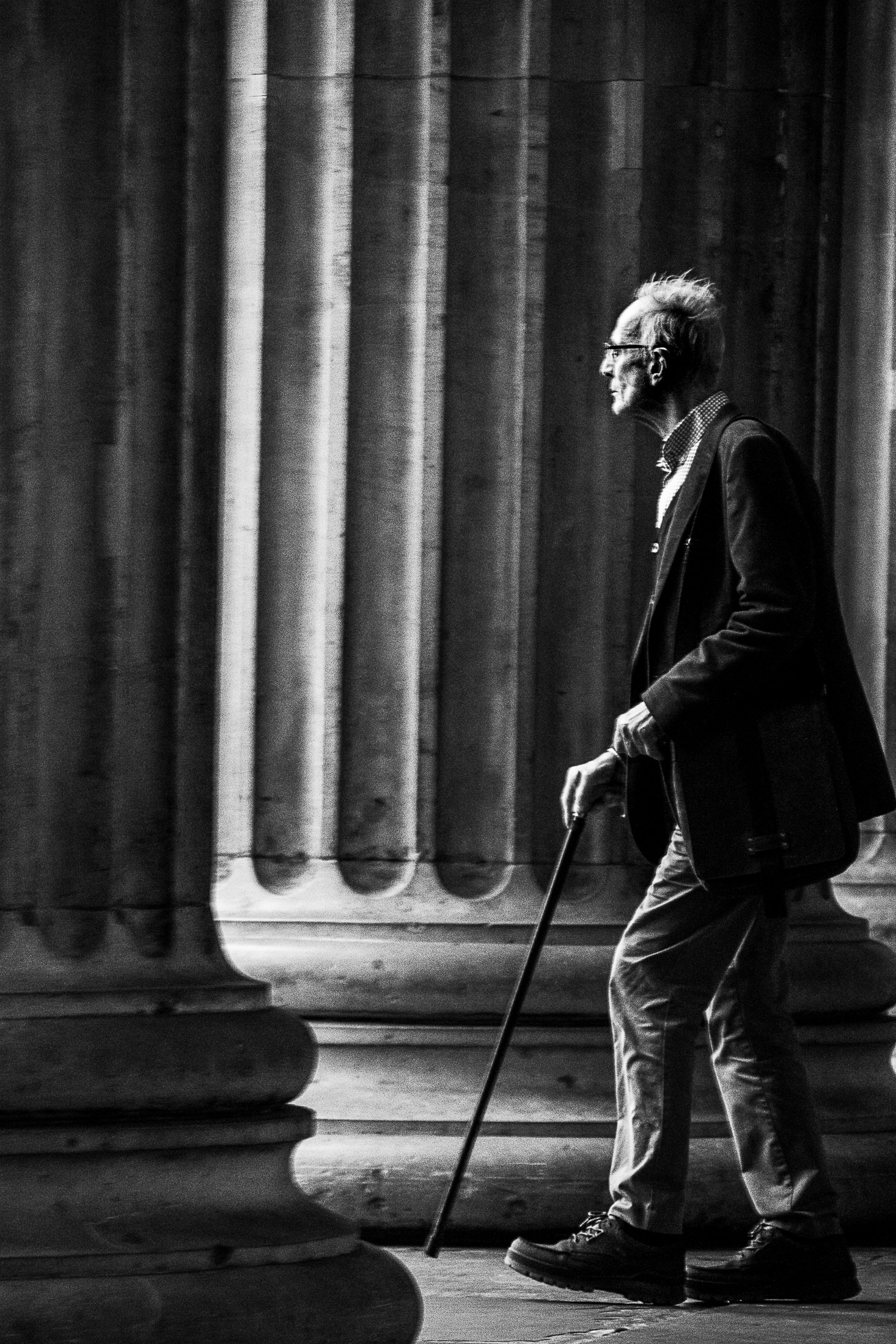 Black and white photograph of an elderly man walking with a cane in front of classical stone columns.