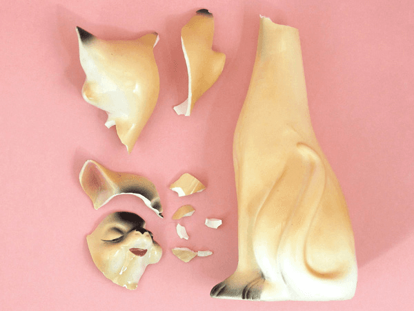 The image shows a broken ceramic object on a pink background. The object is primarily beige or cream-colored and has a smooth, glossy surface. It is broken into several pieces, including one that resembles a face with eyes closed and a smaller piece that looks like a hand. The pieces are scattered around the object, with no visible pattern or order. The pink background provides a stark contrast to the beige object, highlighting its fragility and broken state.