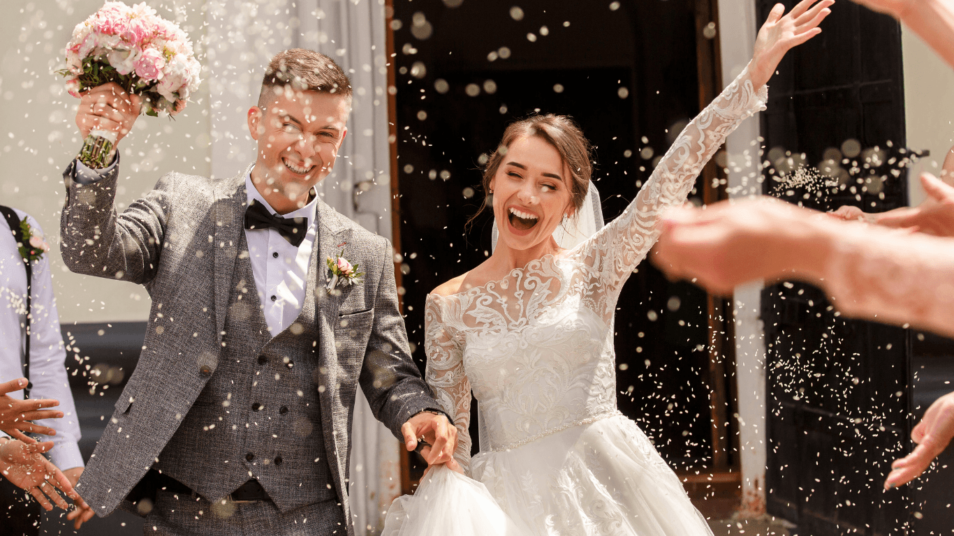 A happy couple in wedding attire celebrates outside, surrounded by guests throwing confetti.