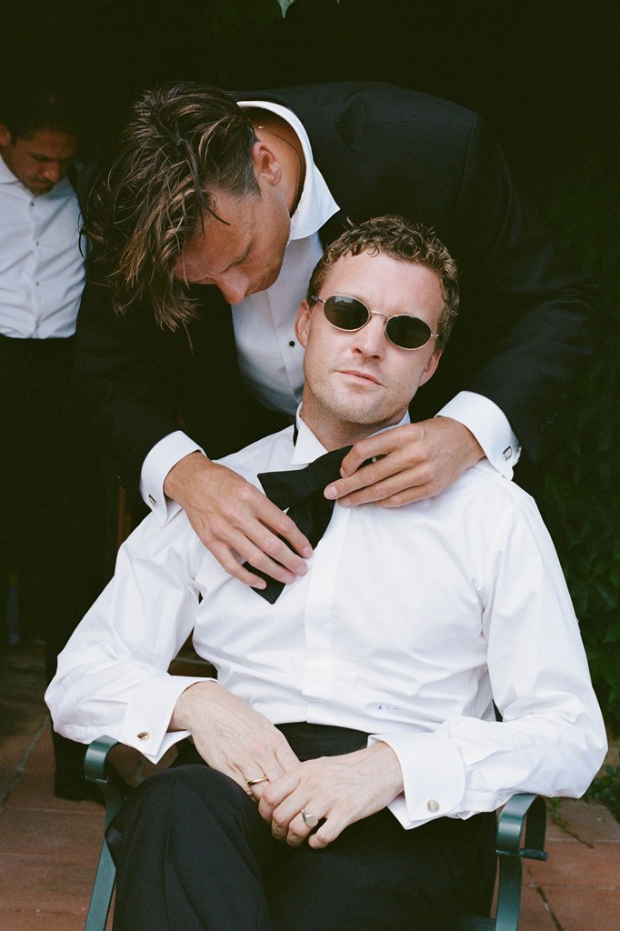 Groom wearing sunglasses being helped into his tuxedo during wedding preparations