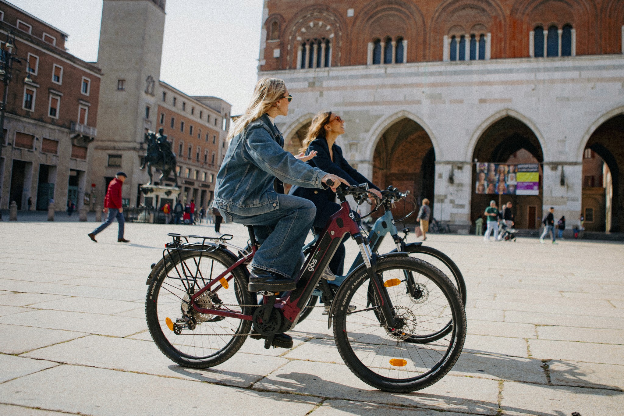 Due donne pedalano su e-bike GBS in Piazza Cavalli a Piacenza, davanti al Palazzo Gotico