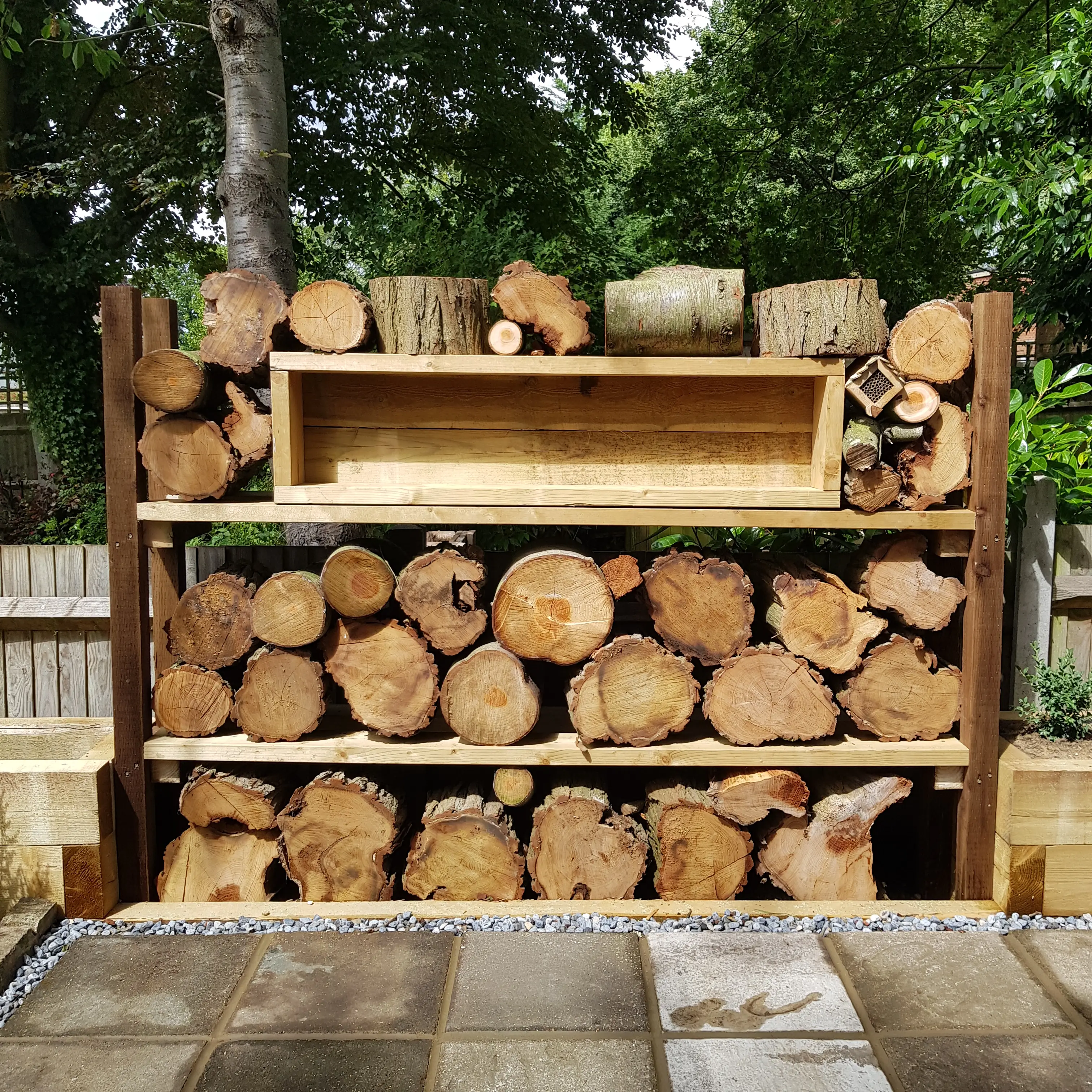 A neatly stacked firewood rack with various sizes of logs, set against a green outdoor backdrop.