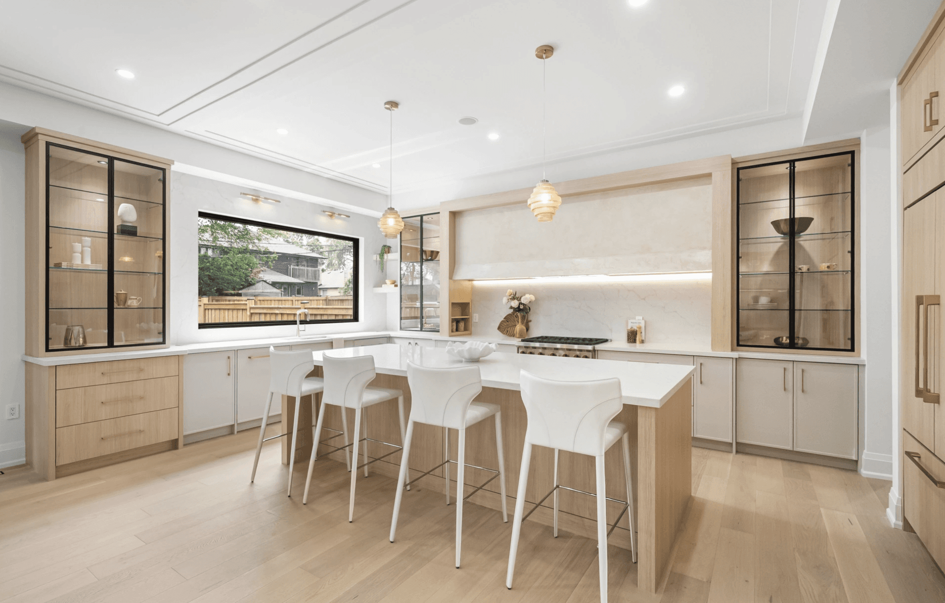 Modern open-concept living room with a large beige sectional, a wall-mounted TV, sleek barstools, wooden accents, and natural light from large windows. Cozy atmosphere.