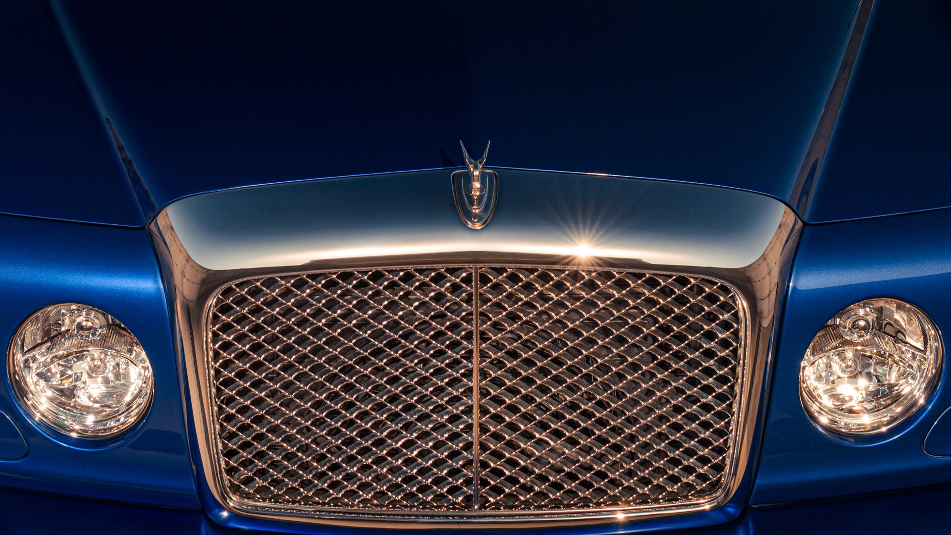 Front detail of a Bentley Brooklands in Moroccan Blue showing the chrome matrix grille, the Flying B bonnet mascot, and round headlamps lit against the deep blue paint, with a sunburst flare across the bonnet.