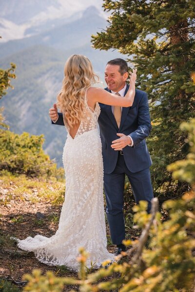 The bride reaches toward a smiling man in a blue suit beside the mountain overlook.