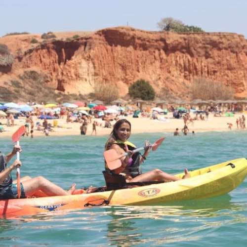兩個人在擠滿海灘的海洋裡划皮艇，周圍有五顏六色的陽傘，背後是一片紅色的懸崖。