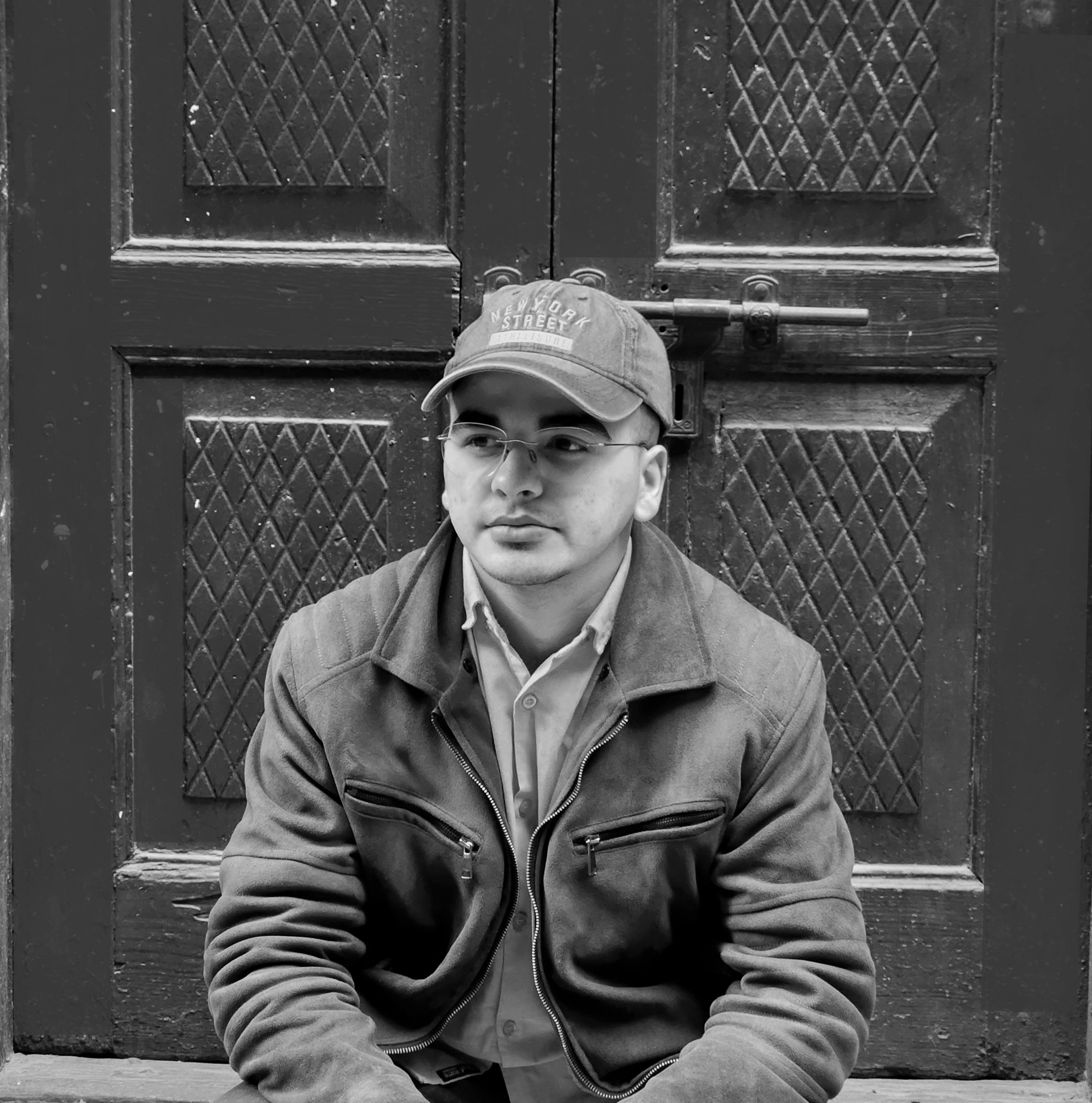 Profile image of Sajid, a digital product designer, wearing a cap and jacket, sitting in front of a textured door.