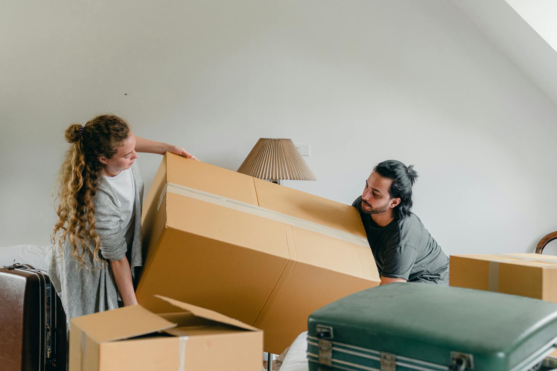A man and a woman are seen struggling to lift and maneuver a large, heavy cardboard moving box. The man has long dark hair tied in a ponytail and is wearing a dark t-shirt, and the woman has long curly blonde hair and is wearing a gray cardigan. A vintage suitcase sits on the floor in the foreground, and a bedside lamp is visible in the background.