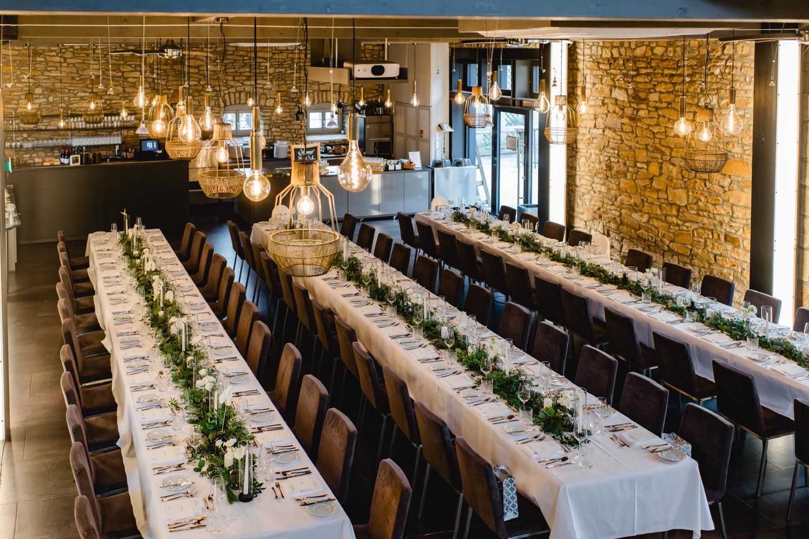 Professionell eingedeckte lange Tafel für ein Hochzeitsevent in einer Location mit Natursteinwänden und moderner Beleuchtung