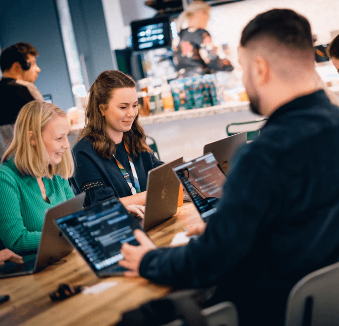 Several people seated around a shared table in a café-style workspace, using laptops and working collaboratively, with a coffee counter visible in the background.