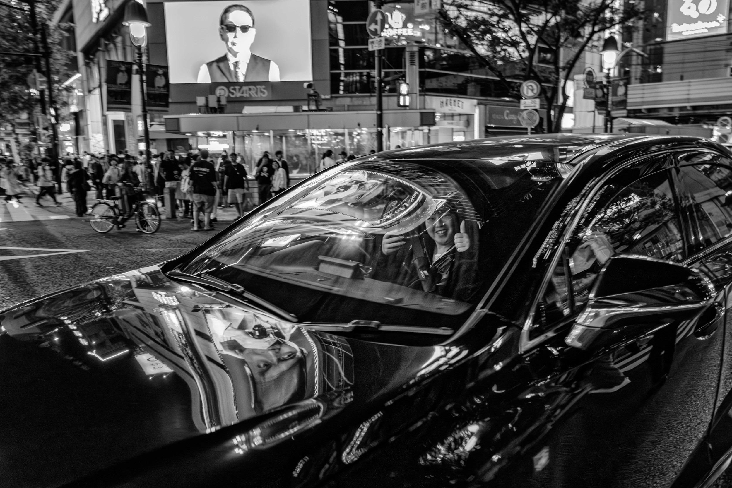 Black and white street photograph of a person giving a thumbs up through a car windshield with reflections of lights.