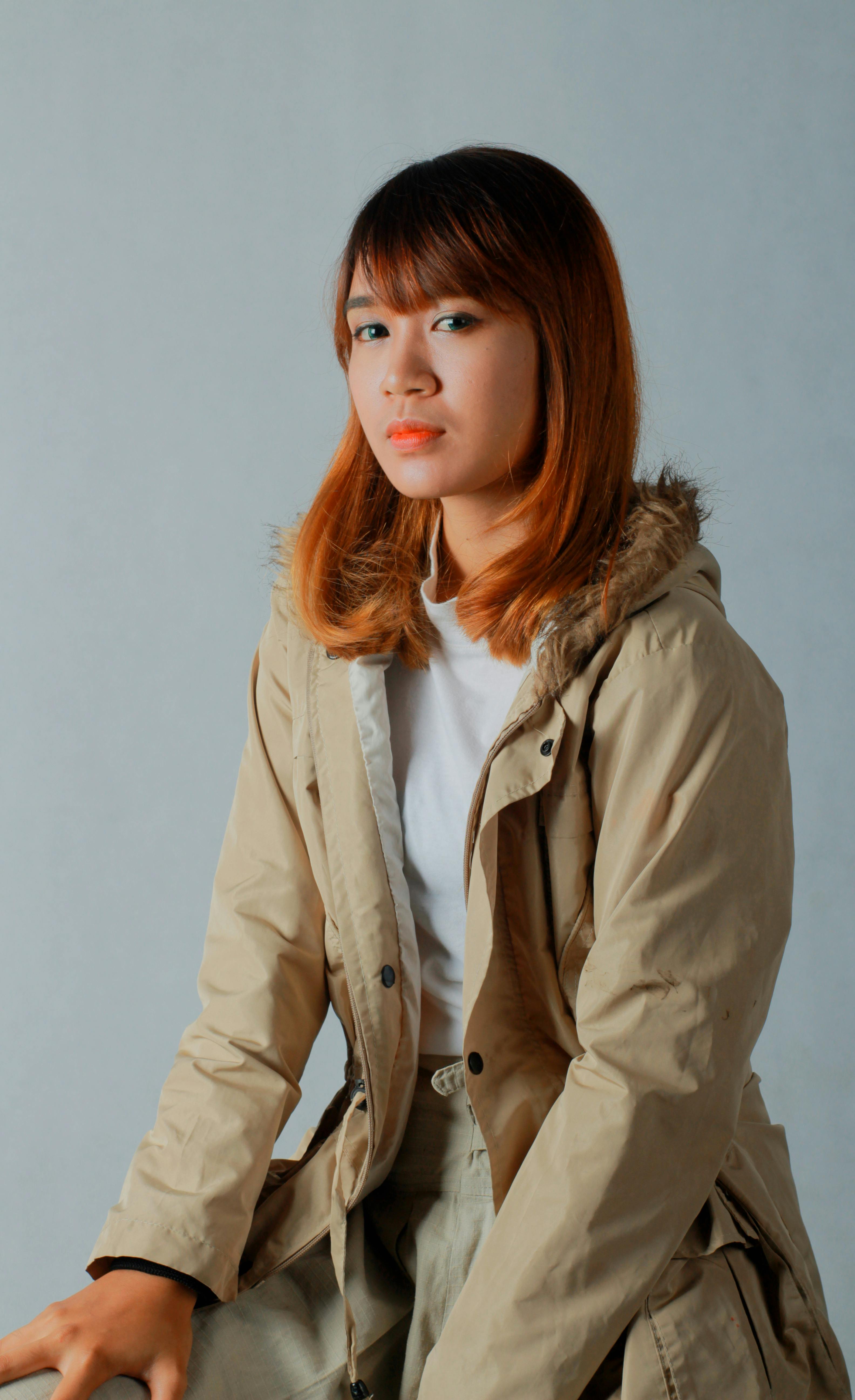 A woman with straight, shoulder-length hair sits against a plain backdrop, wearing a tan coat with a faux fur hood and a white shirt, conveying a calm, thoughtful expression.