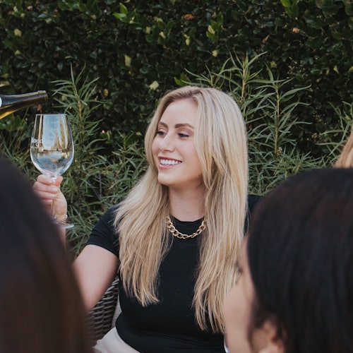 Una mujer con largo cabello rubio sonríe y levanta una copa de vino mientras alguien le sirve vino. Hay follaje verde de fondo.