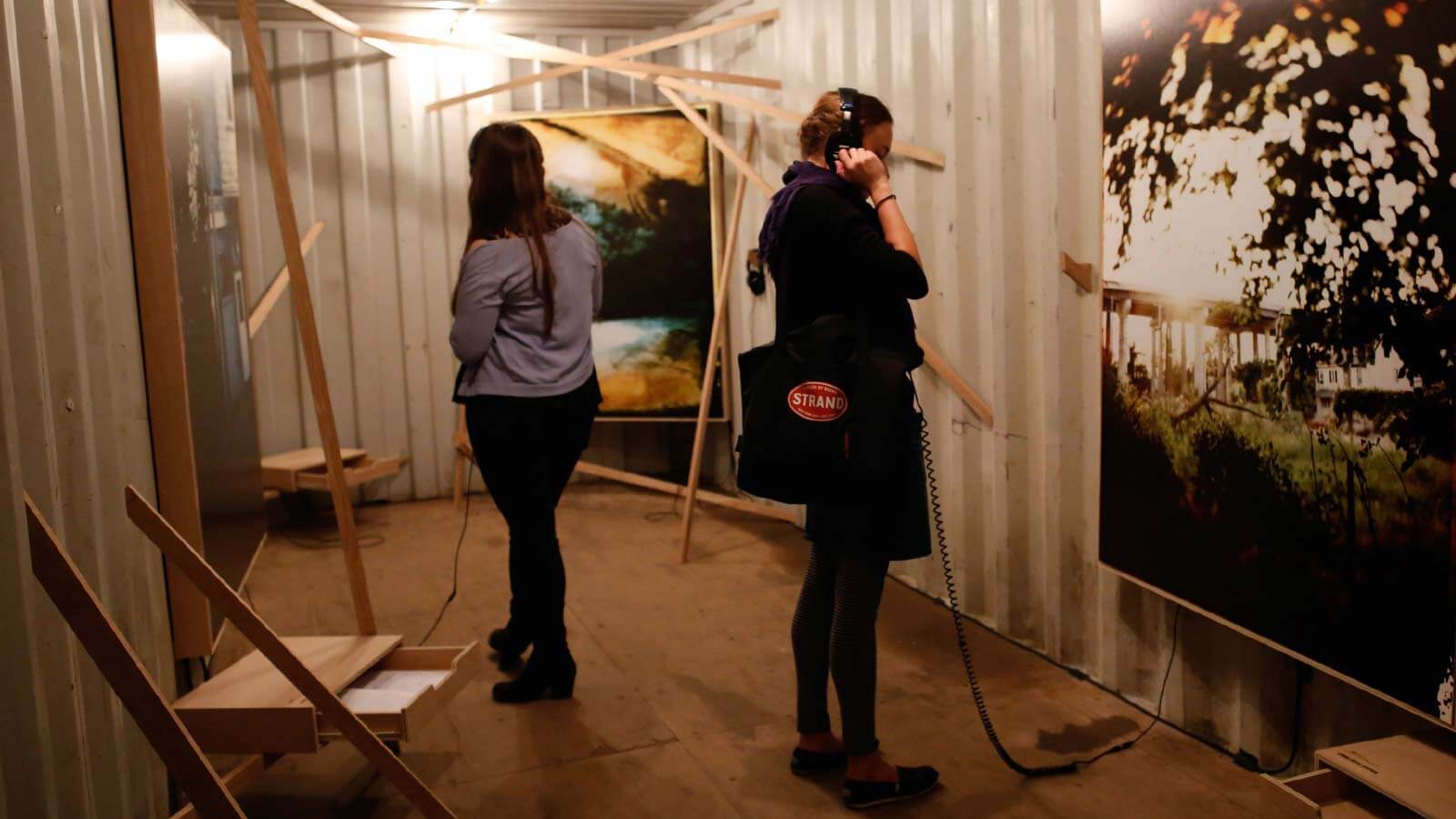Two visitors exploring an immersive art installation with photographs mounted on white shipping container walls and wooden frames