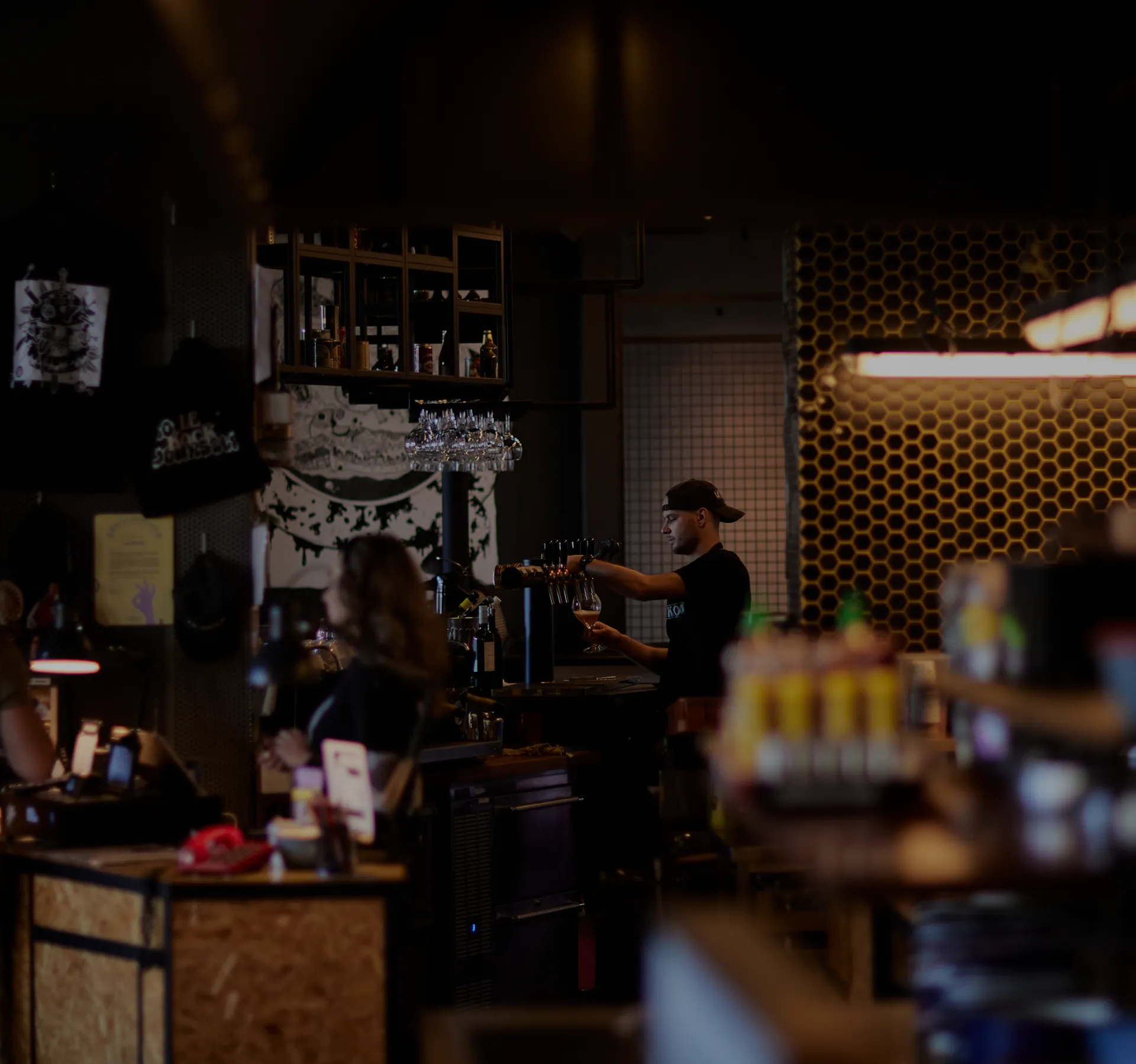 Dimly lit bar interior with a bartender preparing a drink behind the counter, blurred guests in the foreground, shelves of bottles, hanging glassware, and a glowing honeycomb-patterned wall.