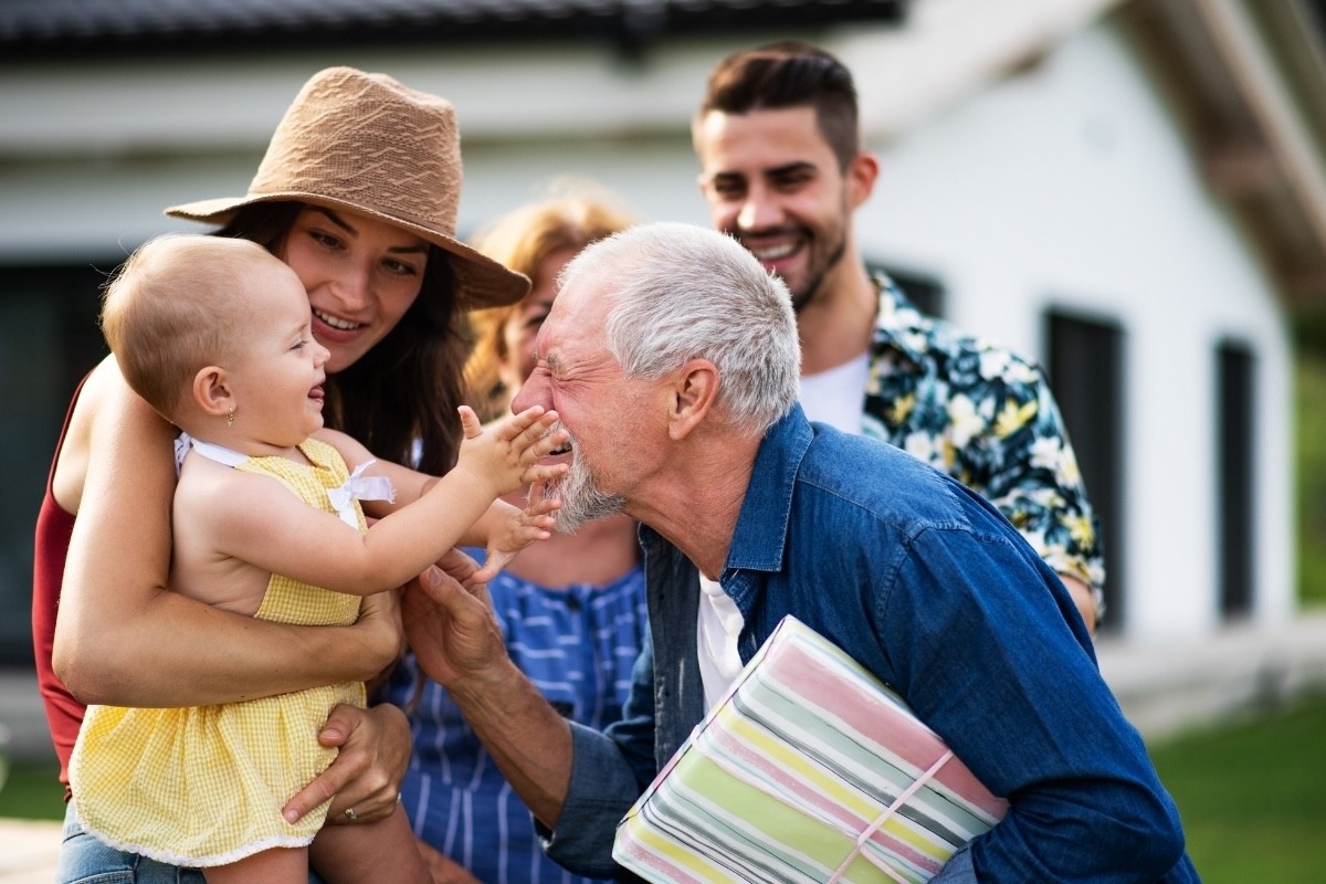 Elderly man greeting family outside before a party and enjoying a playful gesture from his baby granddaughter.