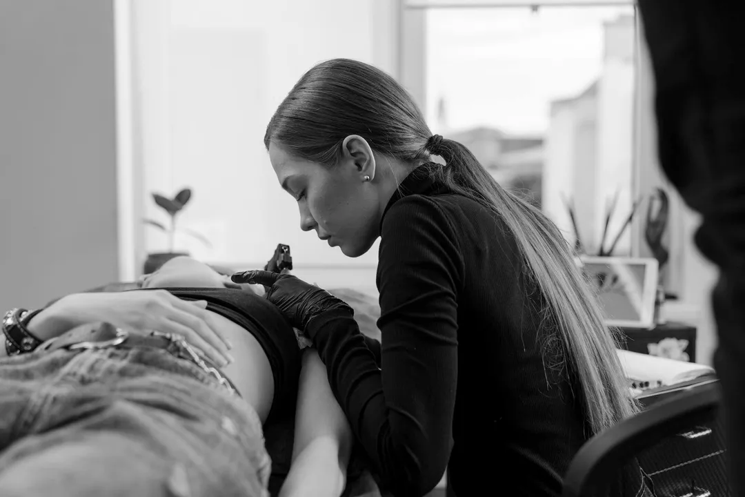 A female tattoo artist focused on tattooing a client's side, with her long hair tied back and wearing black gloves.