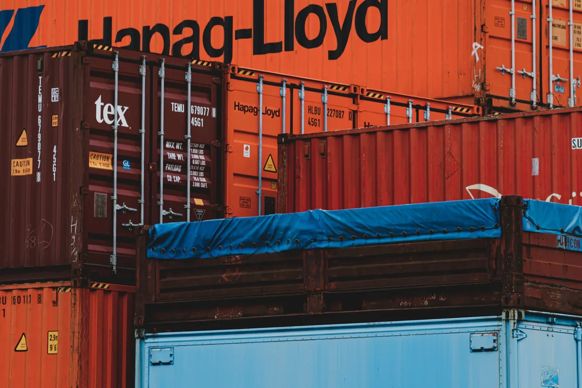 Stacked shipping containers at a UK port awaiting customs clearance, with one open-top container covered by a blue tarpaulin