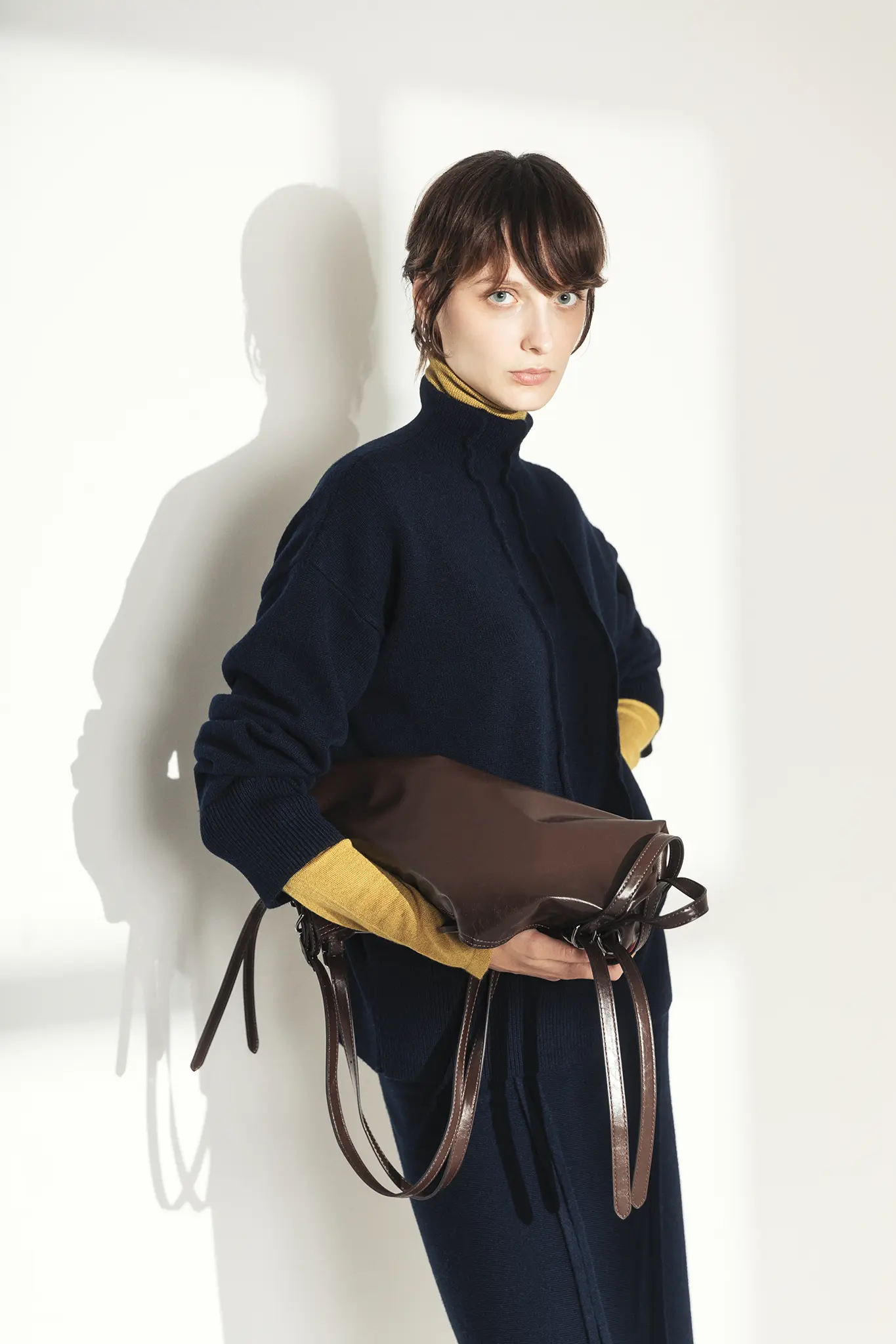 Woman wearing navy cashmere turtleneck layered over mustard yellow undershirt, holding brown leather bag – Aida Barni FW25/26 collection.