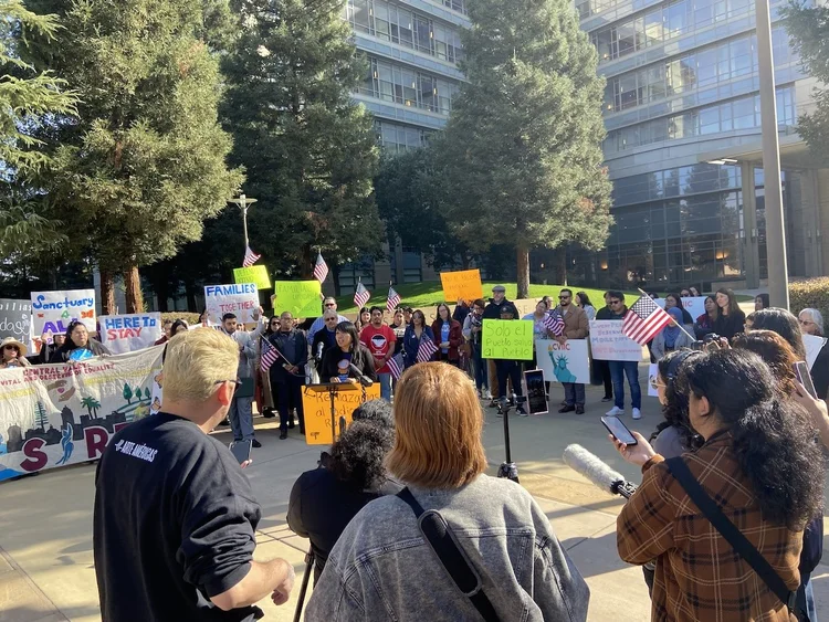 Um grupo diversificado de pessoas se reúne ao ar livre em uma área pública, segurando cartazes e bandeiras enquanto participa de um protesto ou manifestação pacífica, cercado por árvores e prédios modernos de vidro.