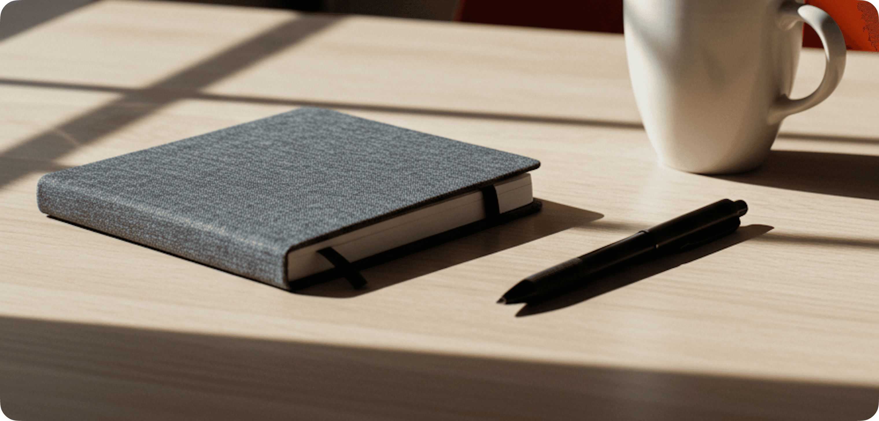 Notebook, pen, and mug on a sunlit wooden table with soft shadows