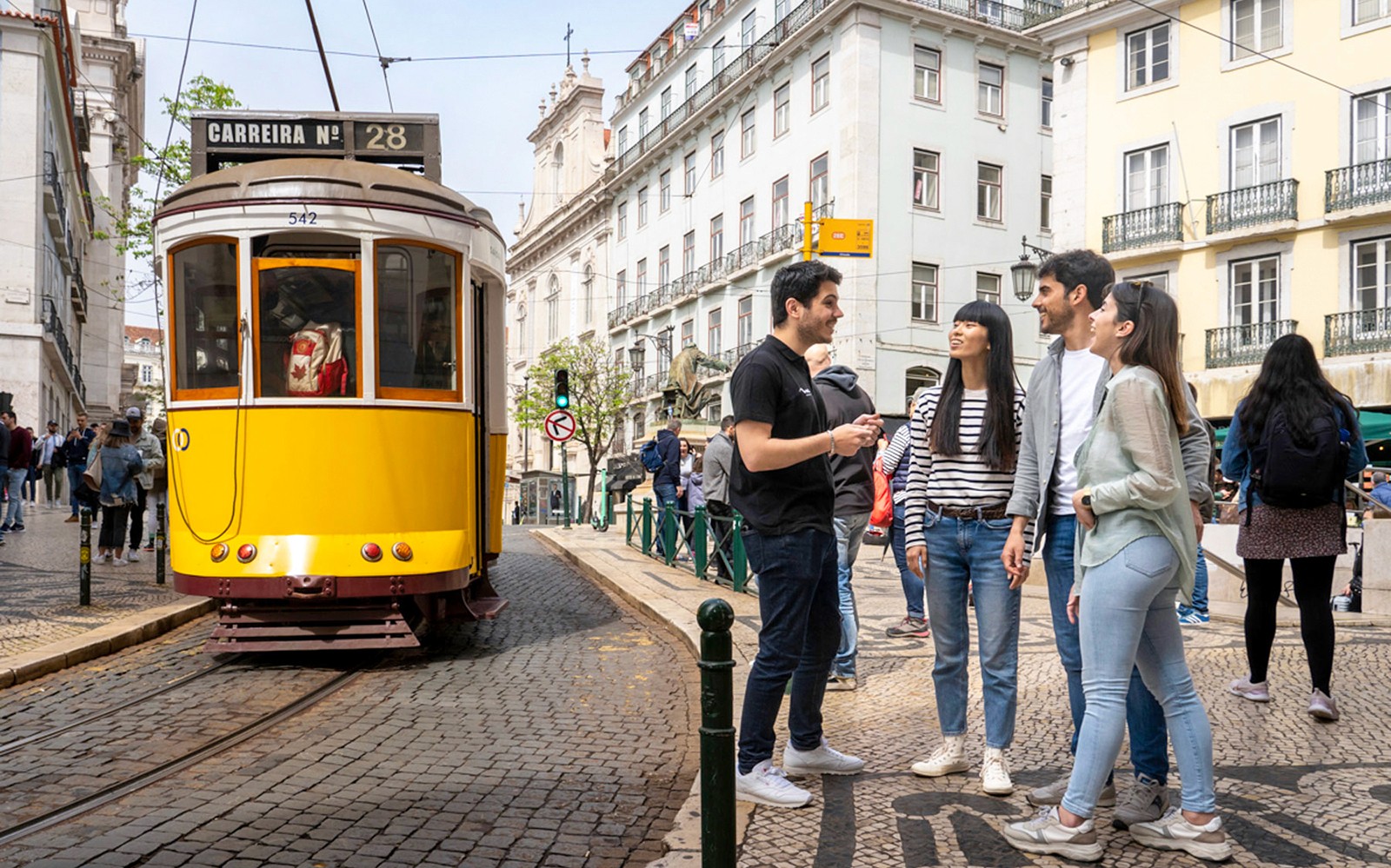 Trem Lisbon 28 melewati kota tua bersejarah dengan sekelompok turis di dekatnya.