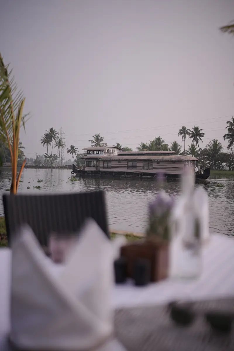 Table set for a Houseboat Wedding celebration at Vedic Village Alleppey, with a traditional Kerala houseboat crossing the backwaters in view